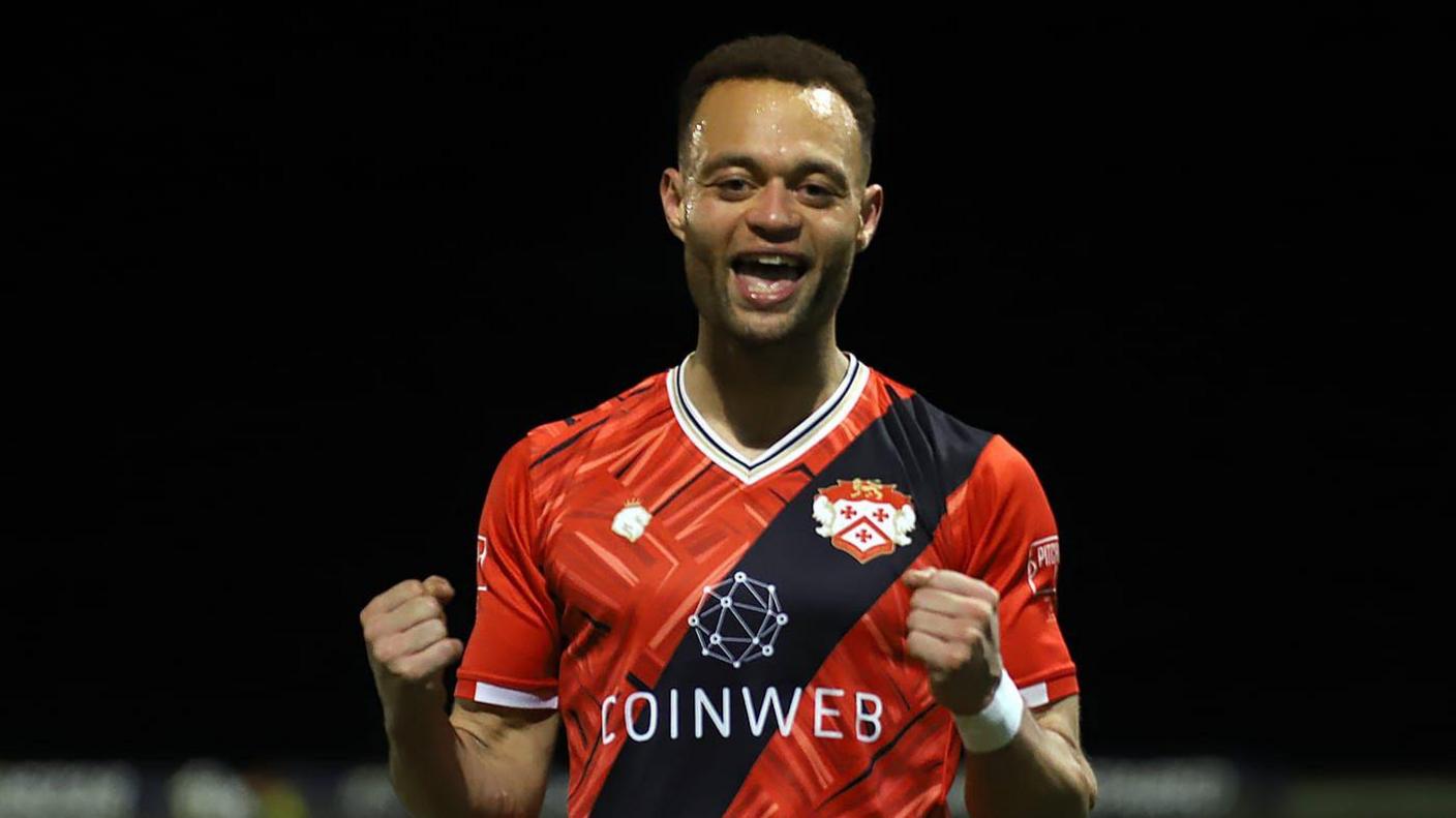 A man with short black hair in a red and black football shirt - has both his fists clenched in triumph. It is dark and he is outside