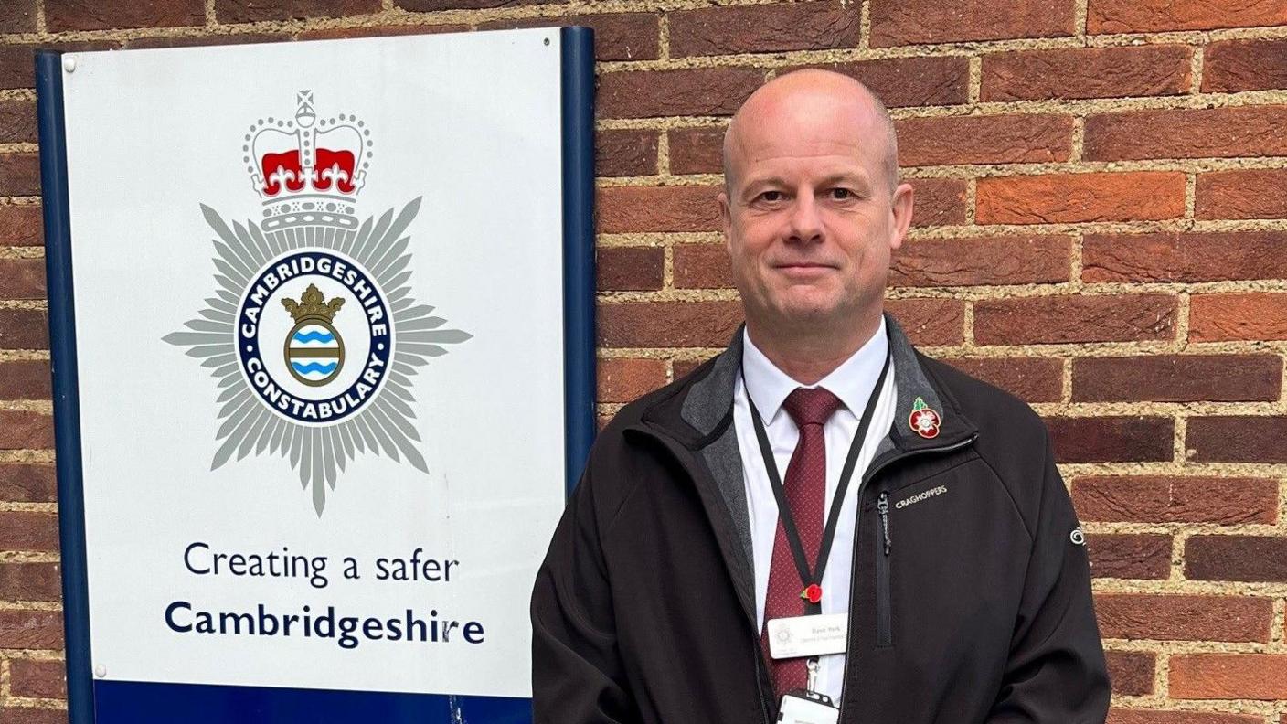 Dave wearing a black jacket and a white shirt and a red tie, standing next to a Cambridgeshire Police signboard smiling for camera.