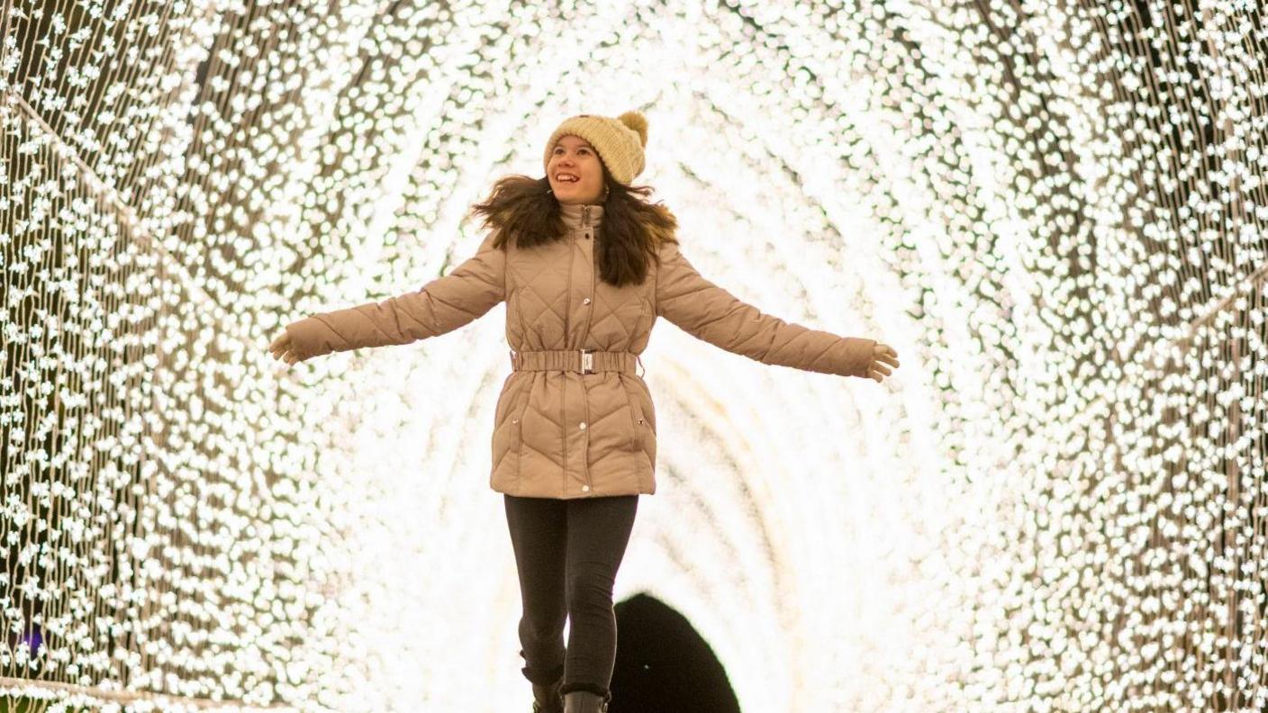 A young teenage girl with long brown hair walks through an illuminated tunnel, made out of strings of fairy lights, at Westonbirt. She is smiling with her arms outstretched and wearing a quilted and belted beige coat, a beige bobble hat, dark leggings, and dark boots. 