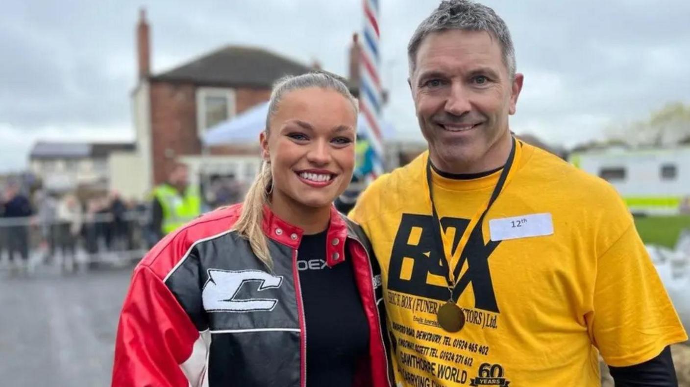 A young woman in a red jacket stands next to a man who is wearing a yellow t-shirt and a medal around his neck.
