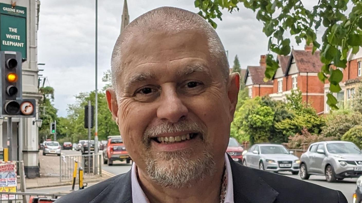 Chris Leggett with short grey hair and beard standing at a road junction