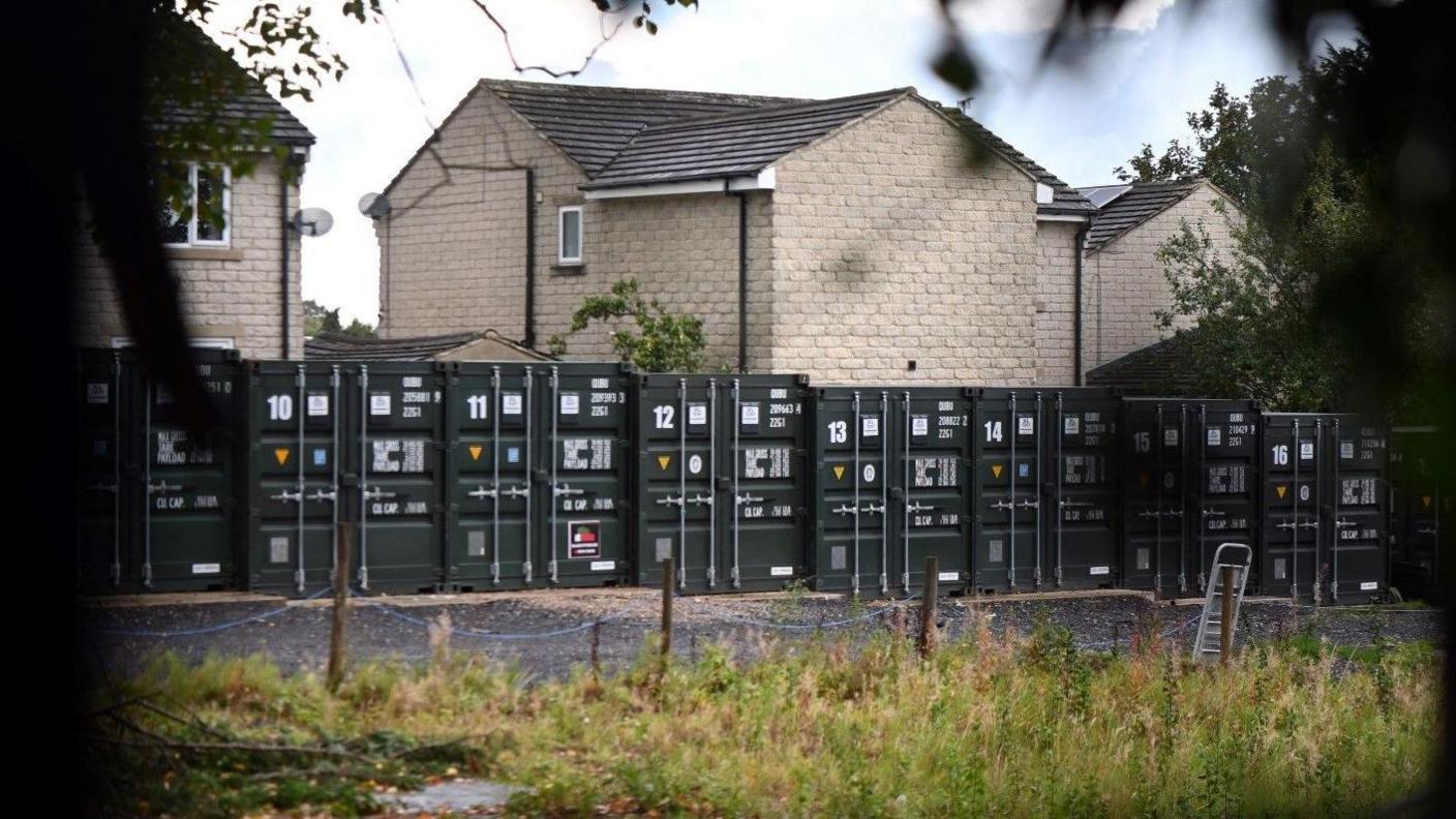A row of metal container units placed side-by-side in a yard in front of houses