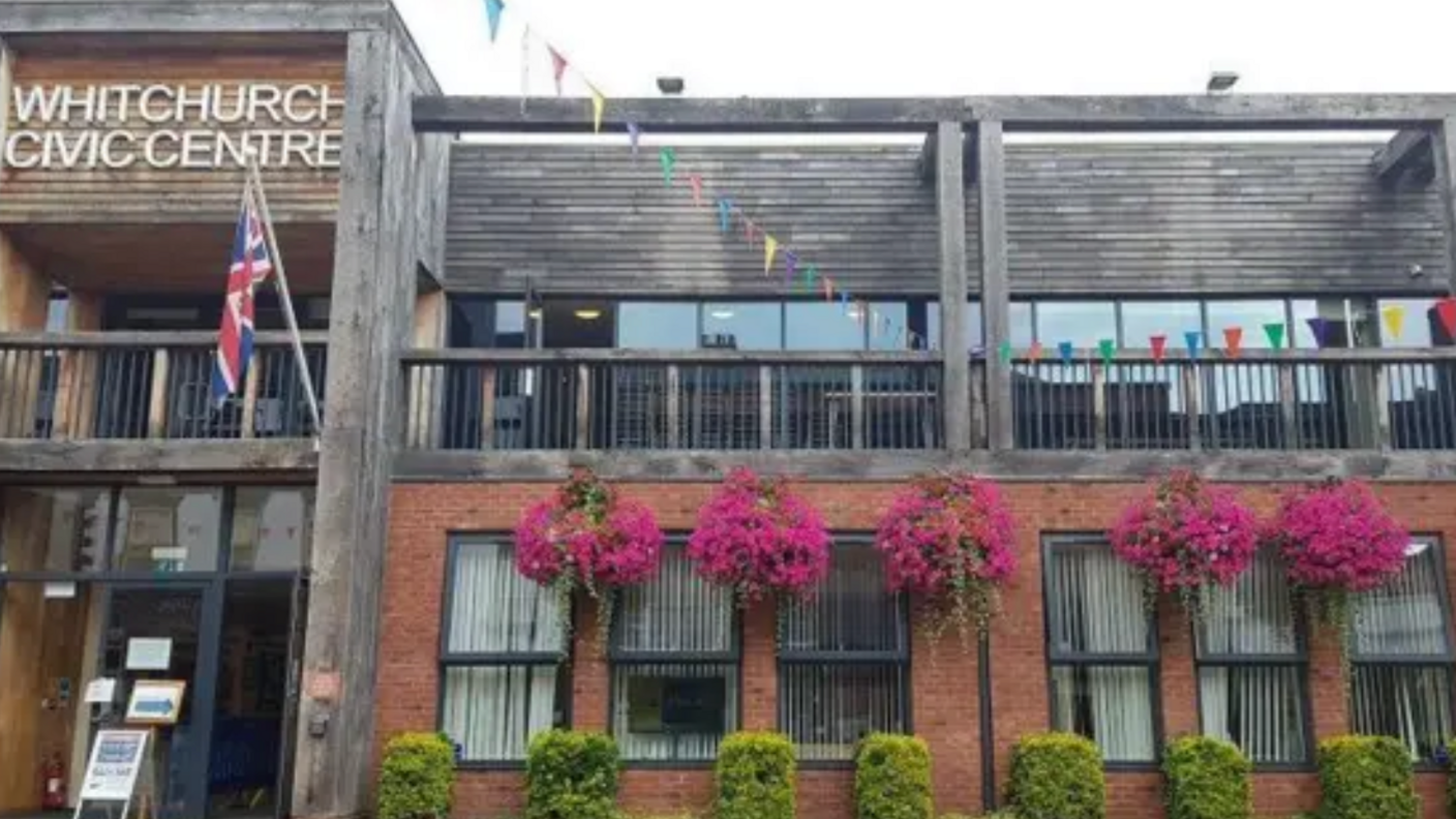 The front of a two storey building. The first floor is made of red brick and has hedges and purple hanging flowers in front of it. The second storey is mostly clad in timber. A sign on the second storey says Whitchurch Civic Centre