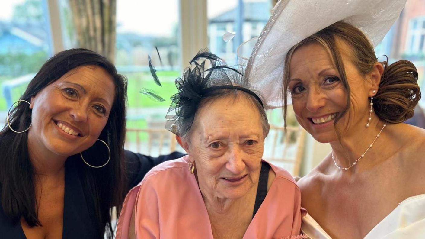 Debbie, Margaret and mother of the bridge Angie smile at the camera while sitting next to each other. Debbie has long dark hair and is wearing large hooped earrings. Angie is wearing a white dress with a large white hat. She has strawberry blonde hair which is tied back.