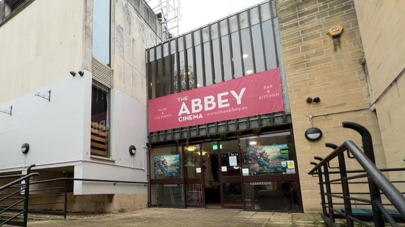 The front facade of abbey hall, including wide steps up to a large glass set of doors, with a red sign saying Abbey Cinema, in white writing, above it.