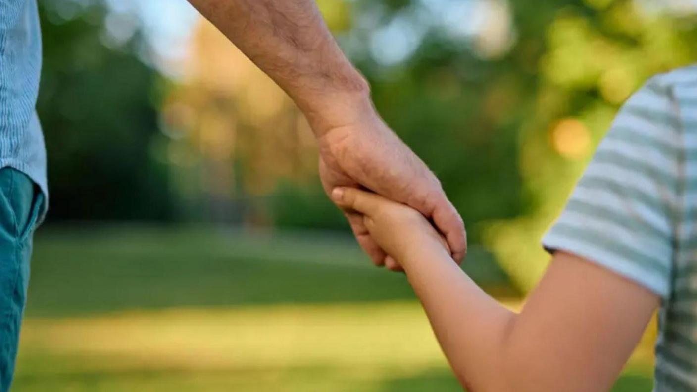 A father and son holding hands as they are walk through a green area. 