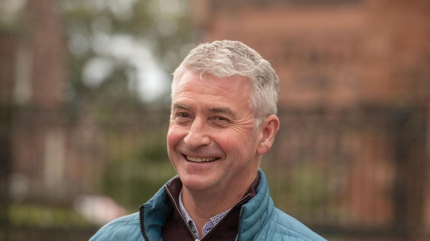 Kevin Beaty. He is wearing a light blue coat with a shirt underneath. He has grey hair and is smiling at the camera. 