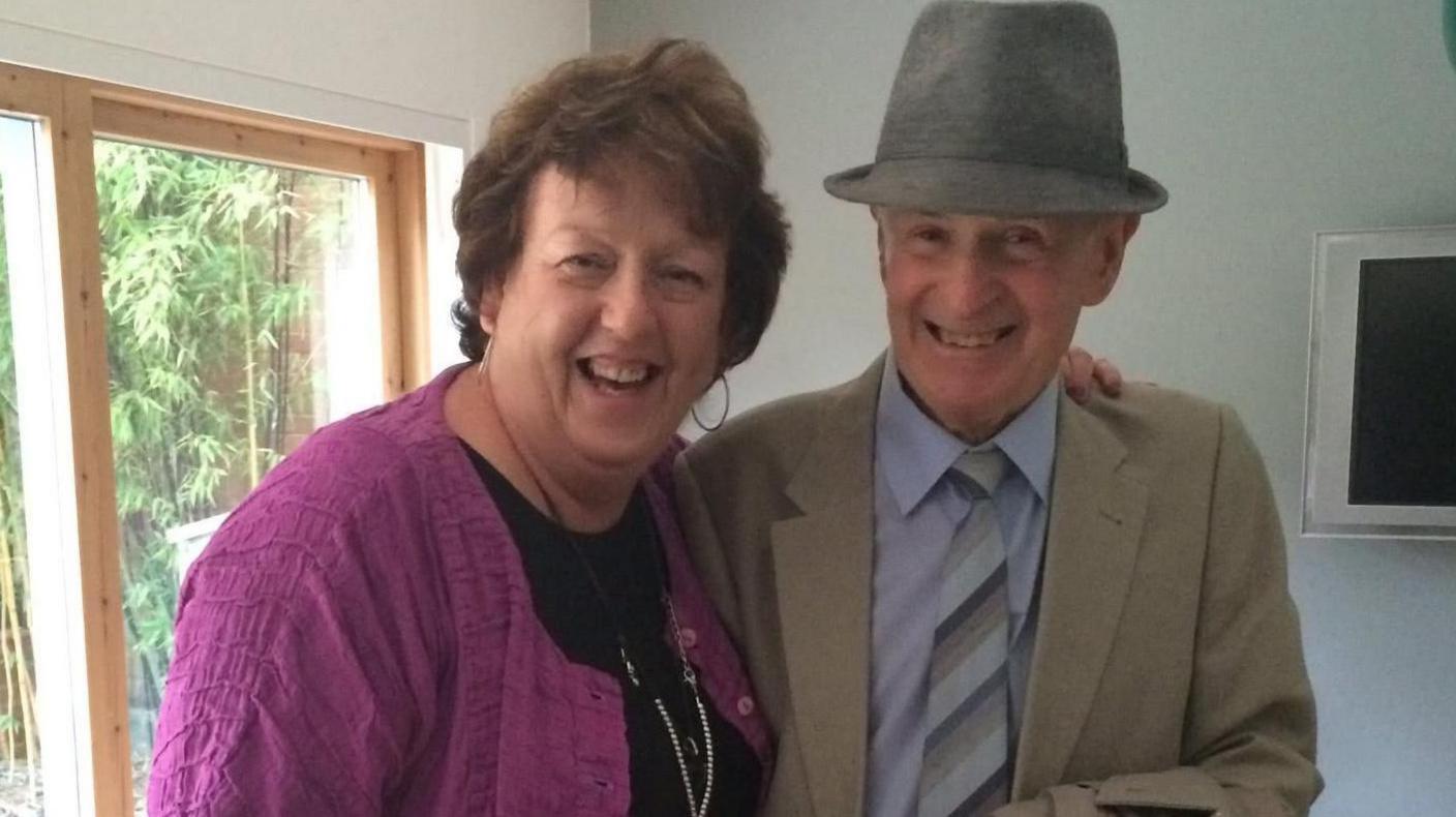 An elderly man smiles (right) for a photo. He is wearing a grey hat and a cream jacket and light blue shirt.  On the left is a woman also smiling with her arm around the man. She is wearing black with a purple cardigan