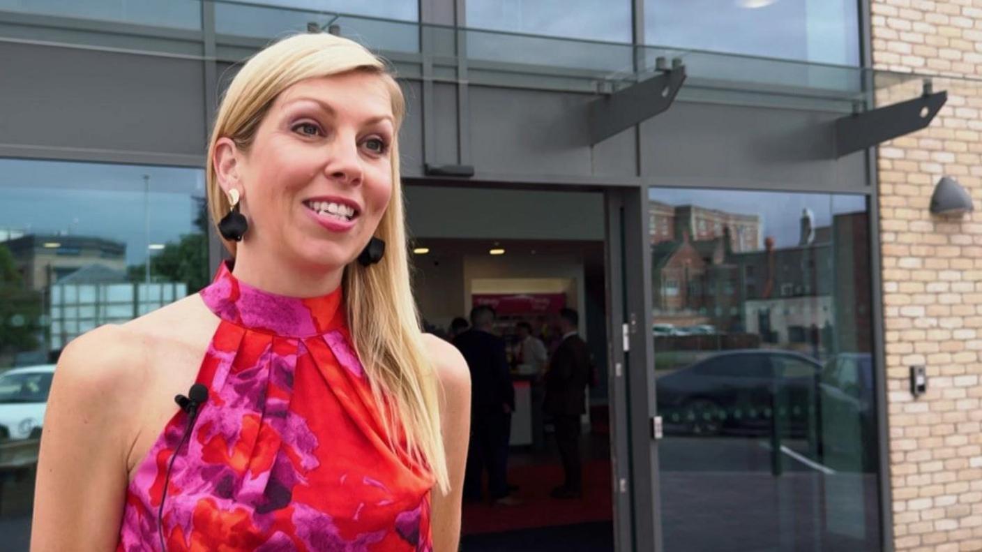A woman with blond hair and wearing a smart red evening dress stands outside a new cinema building with an exterior dominated by large tinted windows.