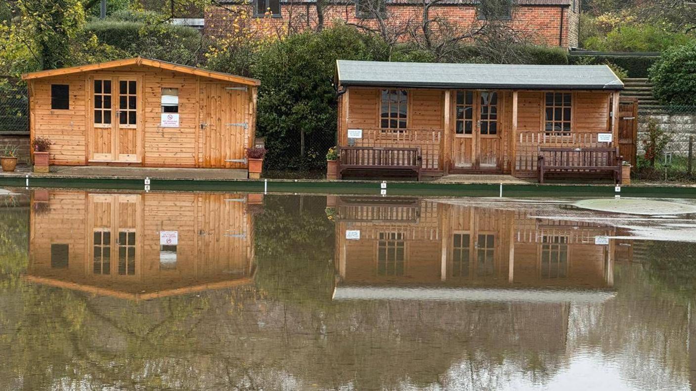 Two brown wooden buildings with water covering the area in front of them. The reflection of the two buildings can be seen in the water.