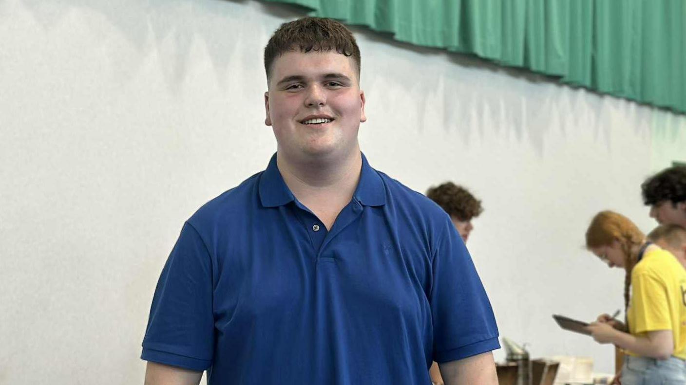 A young, tall man with short dark hair smiles at the camera as he wears a blue polo shirt. Behind him are other young people working at identifying beekeeping equipment.