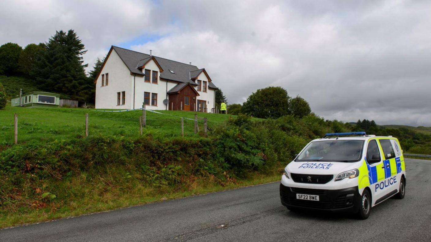 A police van parked below a white-walled detached property. A police officer is near the house.