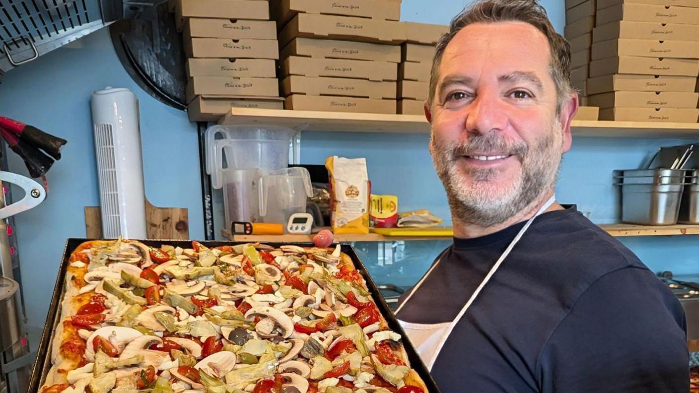 Antonio a man with brown hair and a beard holding a large pizza in a kitchen. The food is covered in peppers and mushrooms.