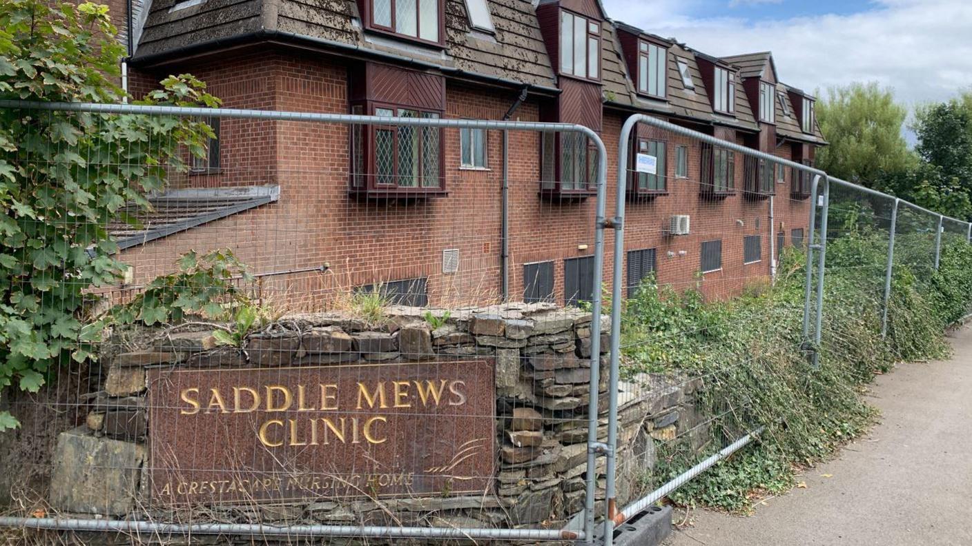 Metal fencing surrounding a stone wall overgrown with foliage with a stone sign engraved with Saddle Mews Clinic. A red brick building is behind with dark brown dormers.