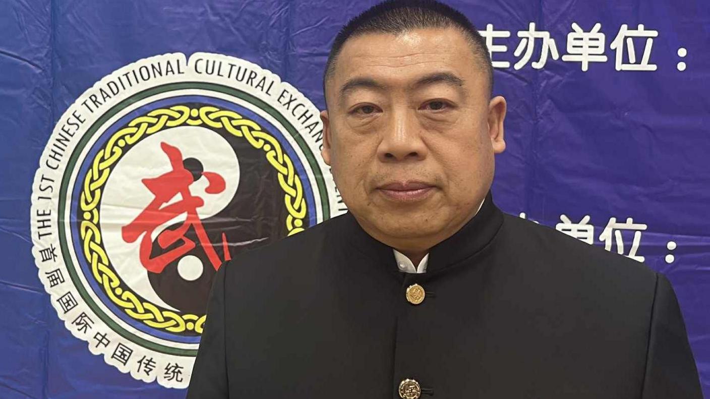 Picture features a man in black traditional Chinese martial arts clothing, standing in front of a blue wall with a logo featuring Chinese writing behind him. There is white writing is featured behind him.