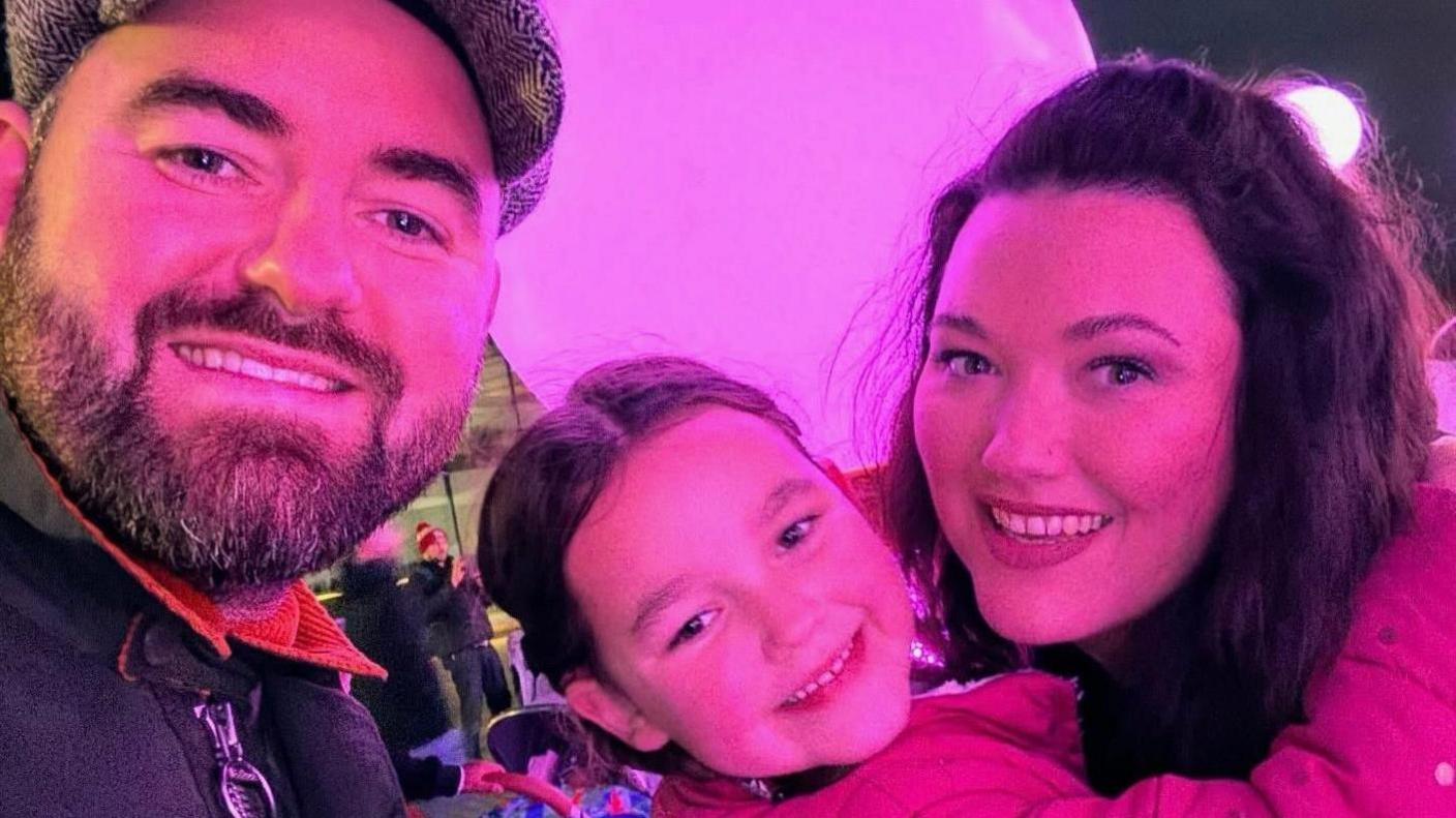 A selfie taken by Ben King as he stands next to his wife Lauren. She is holding their daughter Bebe as they look at the camera.