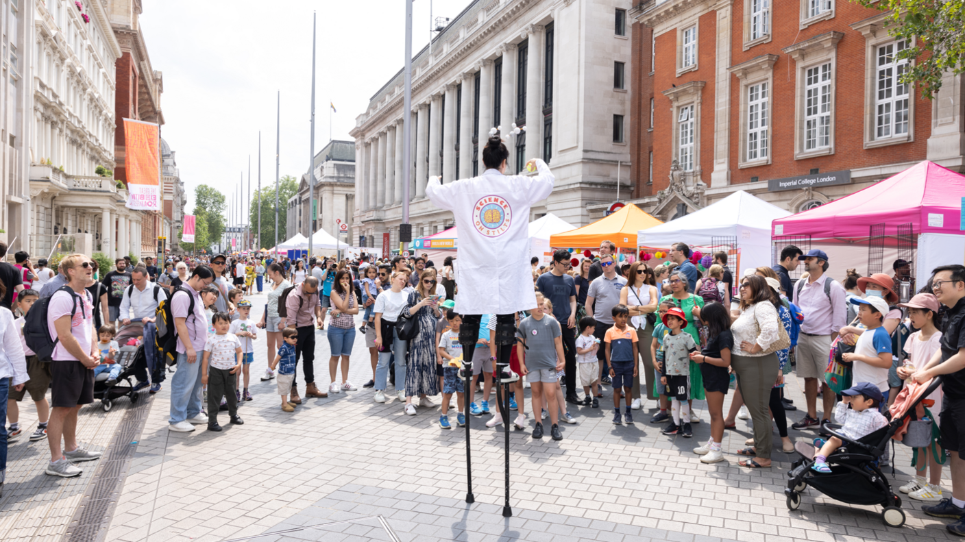 A crowd of people standing on Exhibition Road watching a women standing on stilts wearing a white lab coat. Different colour tents can be seen behind them