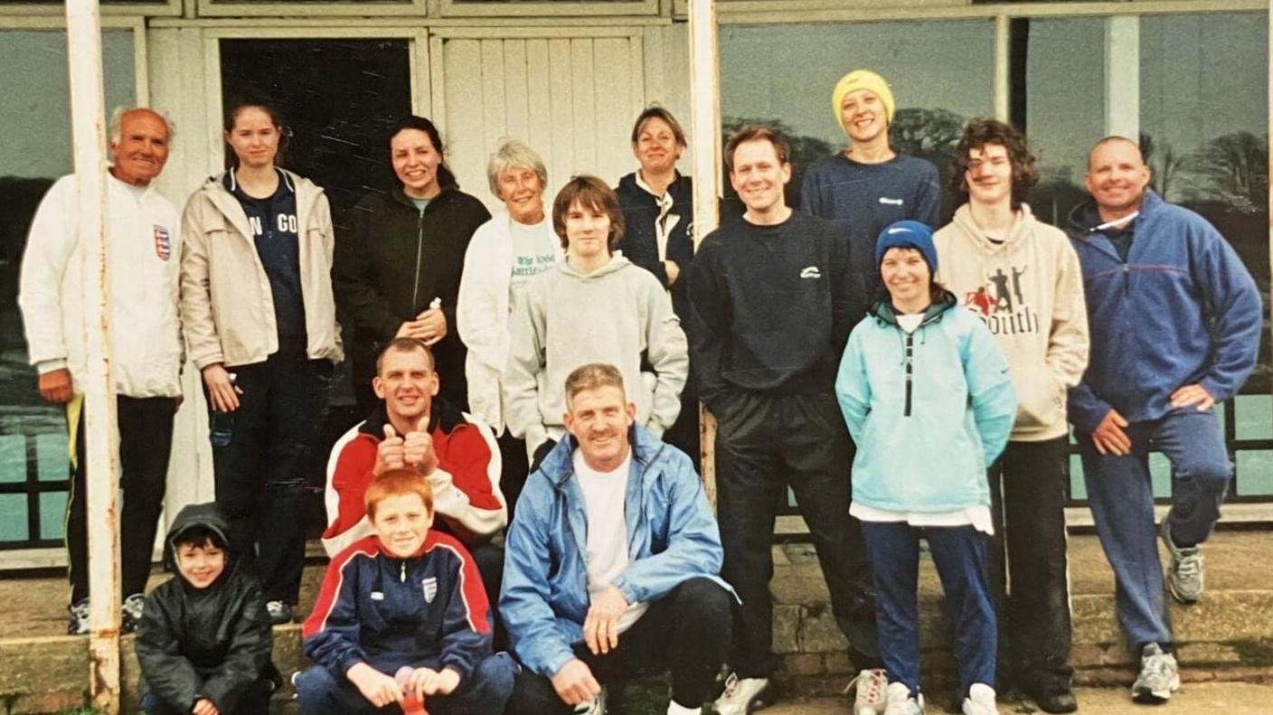 An old photo of a group of people wearing sports clothes standing together outside a sports pavilion. Mr Merfield stands on the far left of the ground wearing a white jumper and black sports trousers. The group smile at the camera. 