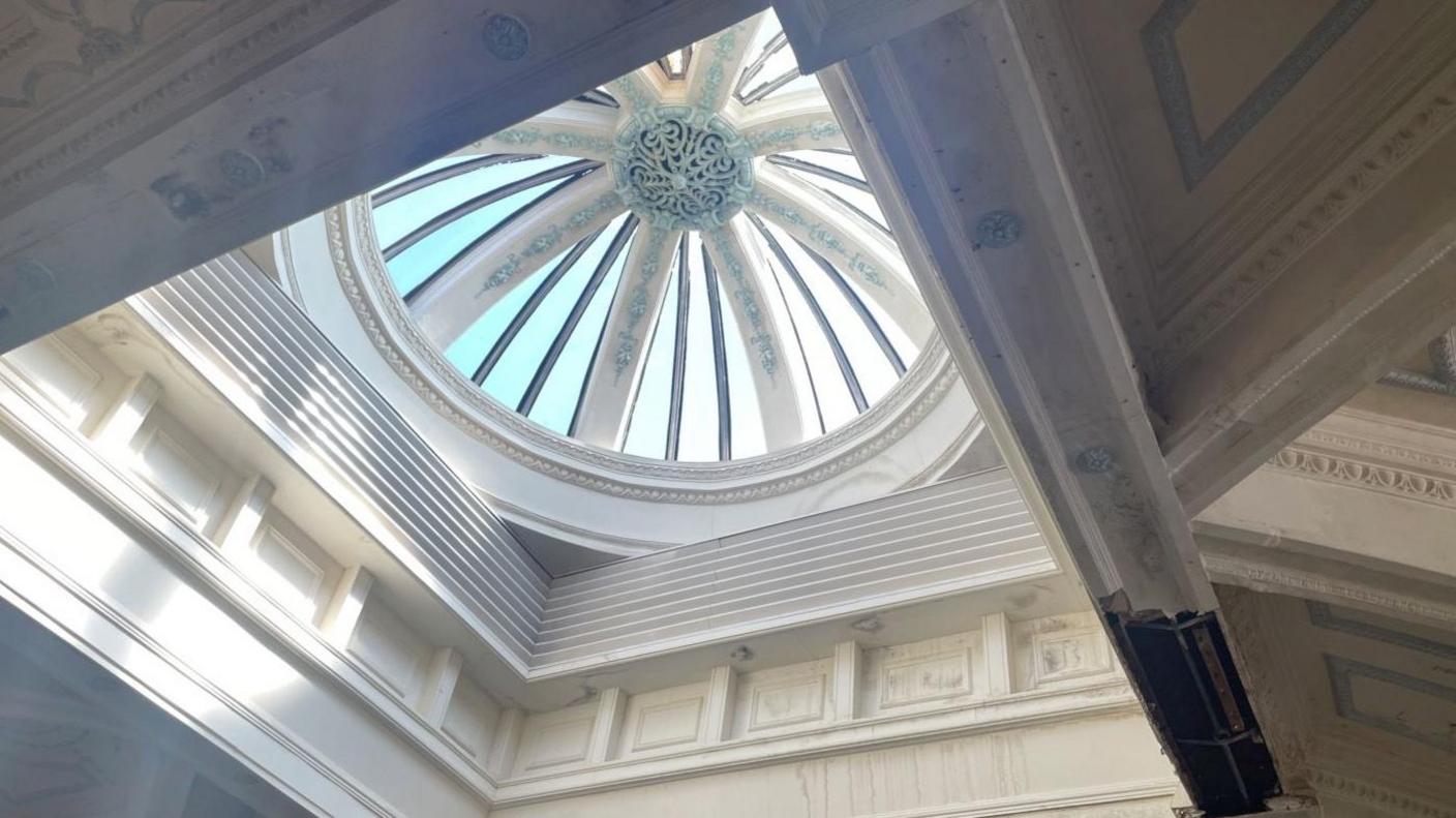 Light shines through a domed window. Next to it a hole has appeared revealing the rotten wood underneath where water has got into the roof.