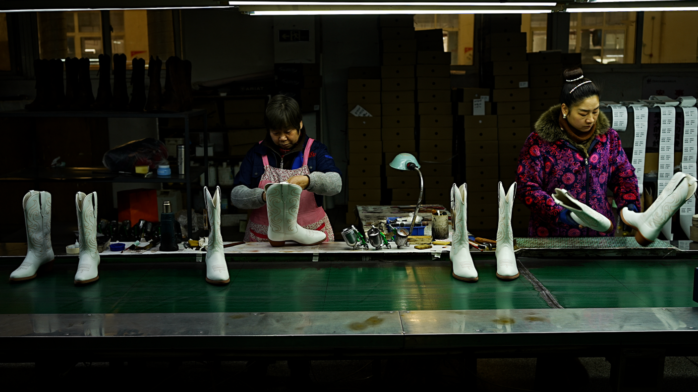 Two female workers making white cowboy boots, which are sitting on a green travellator.  The women are dressed warmly in coats and thick jumpers, while most of the factory behind them appears to be in darkness