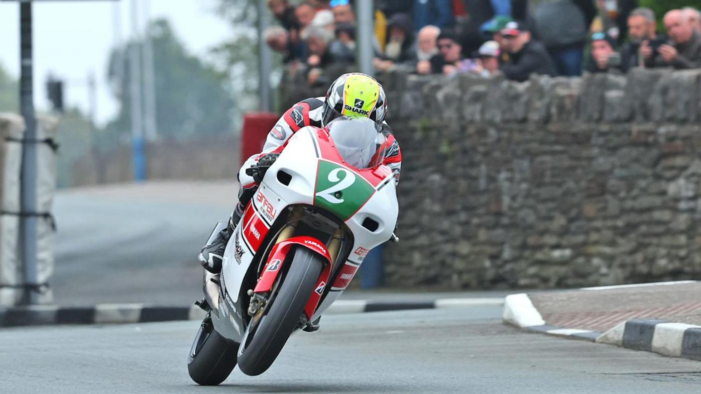 Ian Lougher racing on a white and red bike with a green plate with on the front with the number two on it. He is wearing red, black and white leathers and a white, black and yellow helmet.