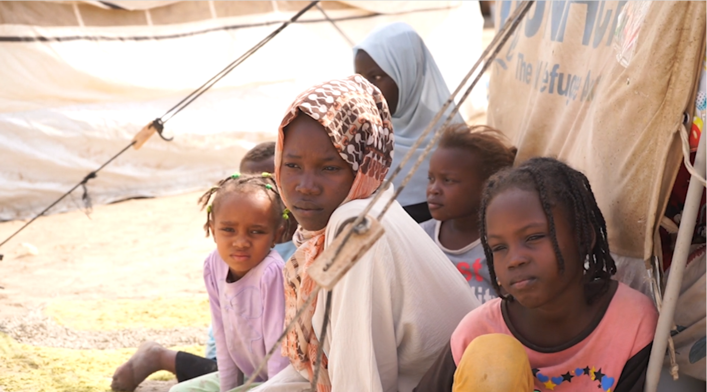 Safaa, a 14-year-old Sudanese girl, is pictured in a refugee camp with five other children. Safaa is wearing a long-sleeved beige outfit and a patterned scarf.