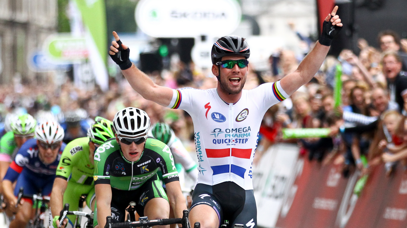 Sir Mark Cavendish cycles over the line with his arms raised and fellow cyclists behind him with crowd cheering in the background 