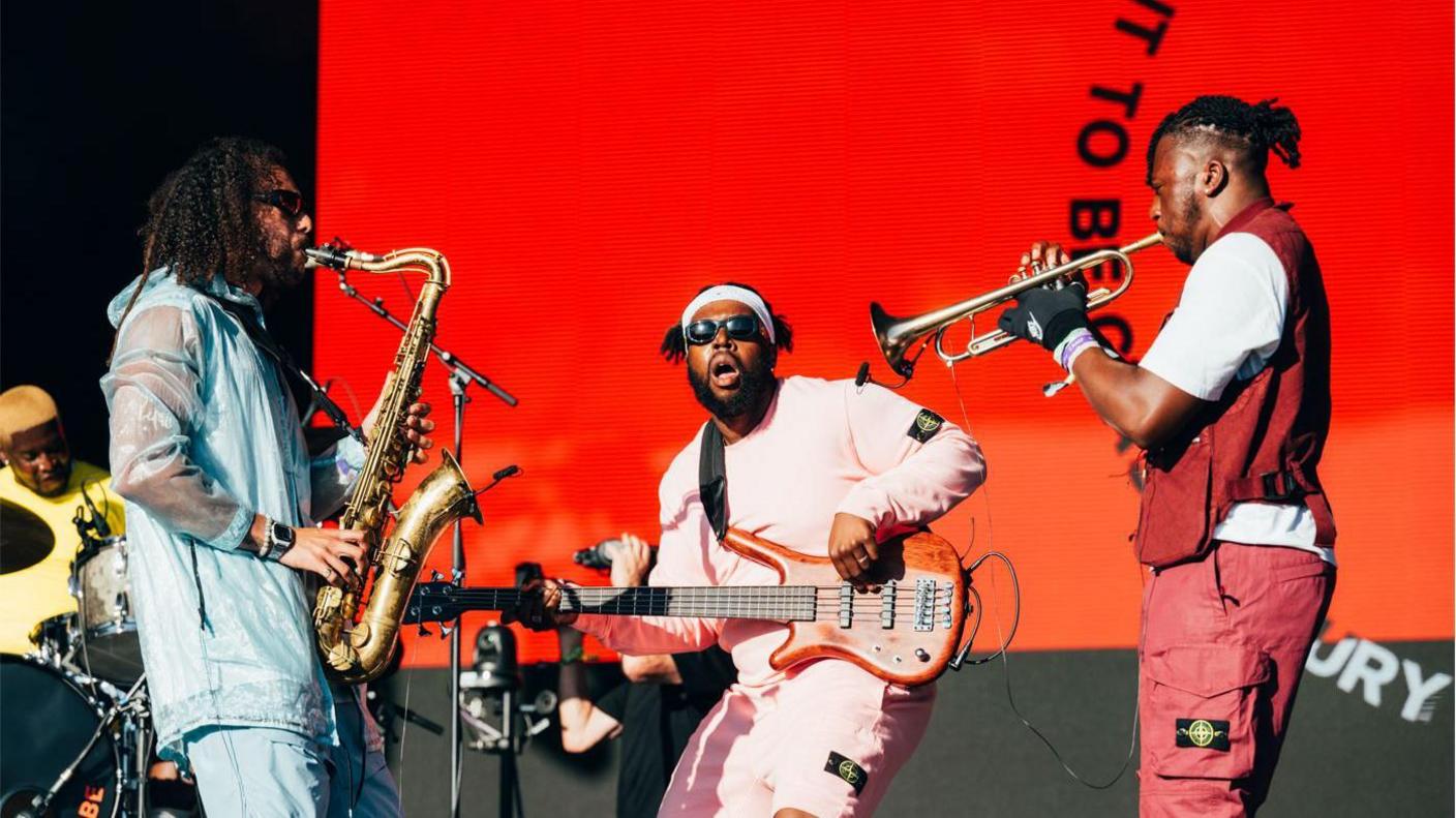 Ezra Collective performing stage at Glastonbury Festival in 2023. A bassist is seen playing in between a trumpeter and a saxophonist, with the drummer seen in the distance. 