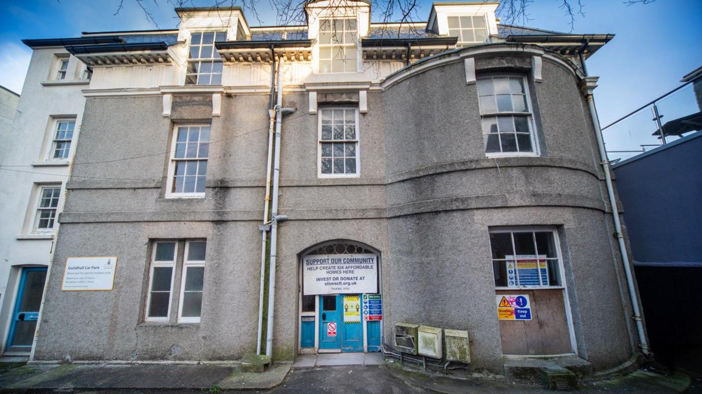 Old Vicarage Flats in St Ives photographed from the ground. The property is three storeys. The exterior is made of dull, grey concrete with some staining beneath the windows. 