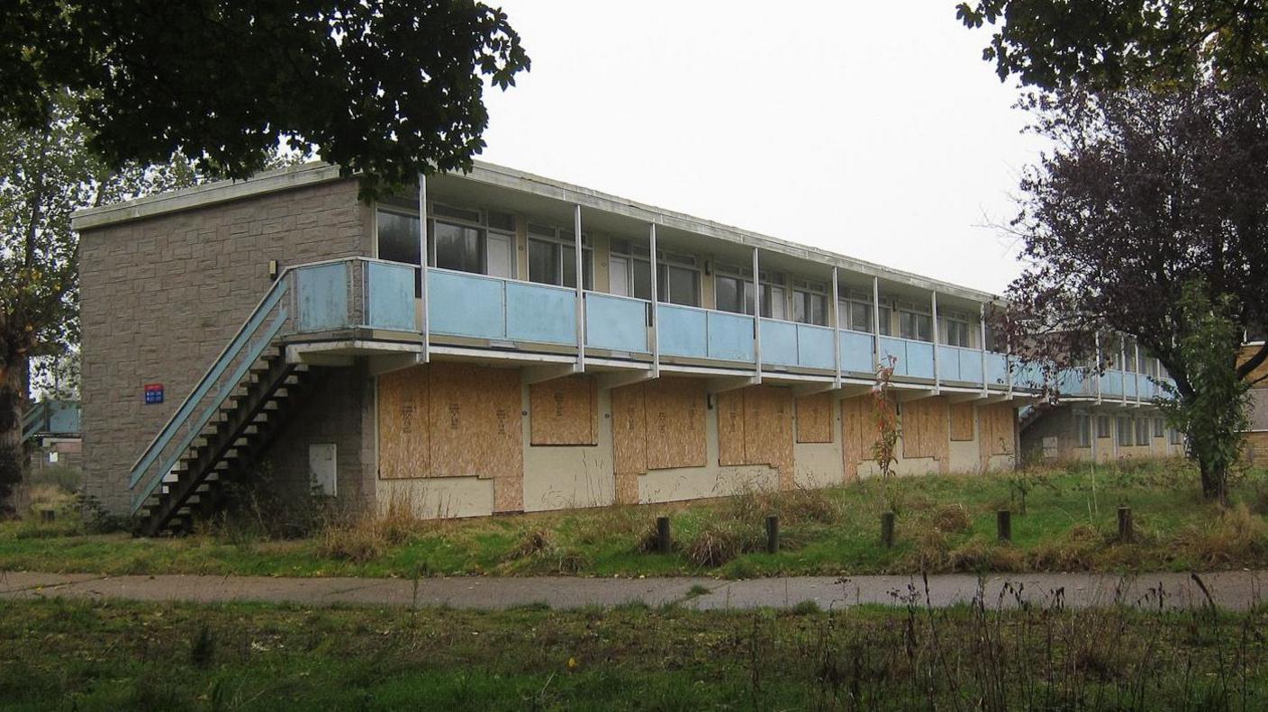 A two-storey building is derelict. The bottom floor's windows are boarded up. A blue painted staircase leads to the second floor. There is grass in the foreground.