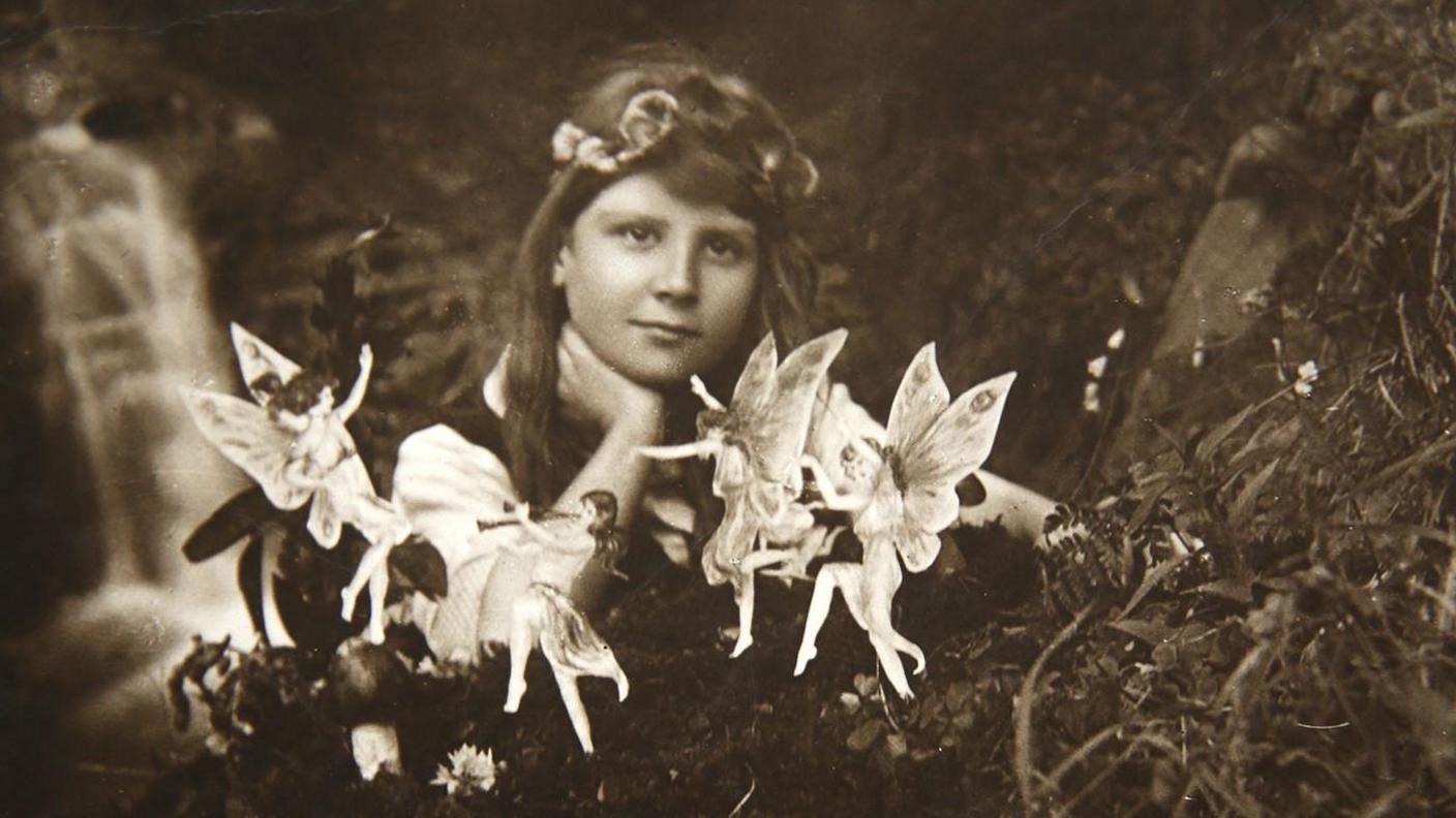 A black and white photo of a young girl with brown hair and a headband resting her hand on her neck and looking at the camera. In front are small dancing fairies. She is sitting outdoors with what appears to be a stream behind her.