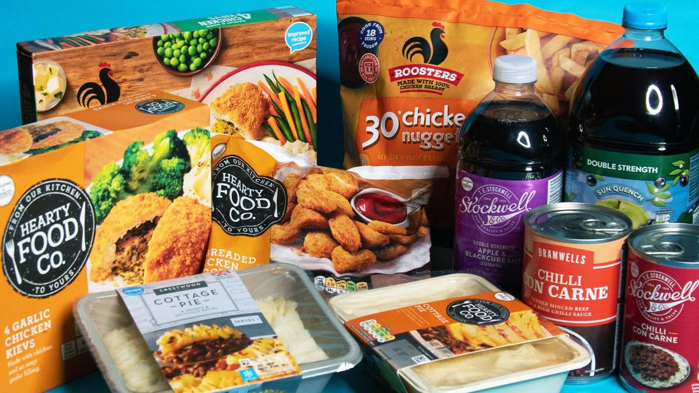 Various food packets from Tesco and Aldi pictured on a blue background. It shows placed next to each other - bottles of squash, tins of chilli con carne, cottage pie ready meal pack, chicken kiev packs and bags of chicken nuggets from each store, with the relevant logo.