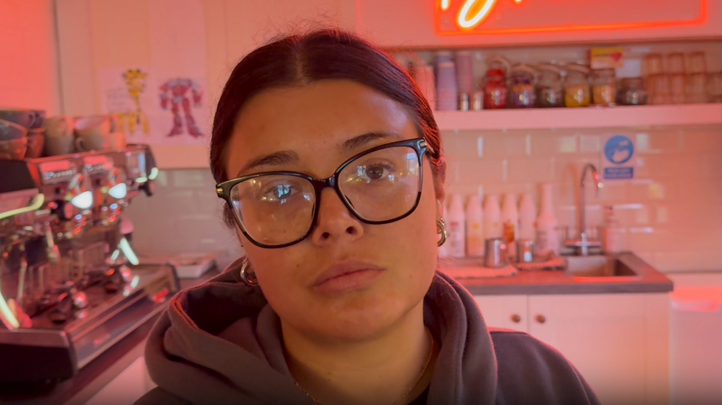 A woman wearing a hoodie stood in a red-lit room. A neon sign sits in the background reading "Blossoms".