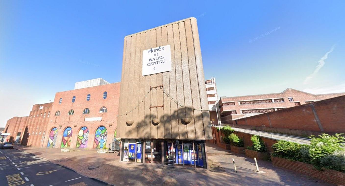 A large building with a ramp at the side of it. There is a sign which reads Prince of Wales Centre above a set of windows and glass doors. A section of the building set back from the entrance has a number of archways which each contain colourful murals.