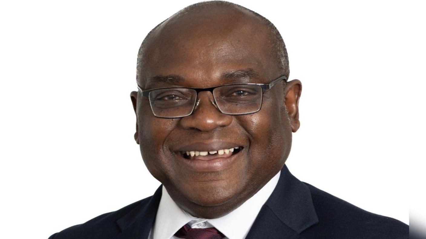 Mr Alexander Gyesi-Appiah - a bald man wearing rectangular glasses smiling with teeth wearing a navy suit white shirt and burgundy tie with a white background