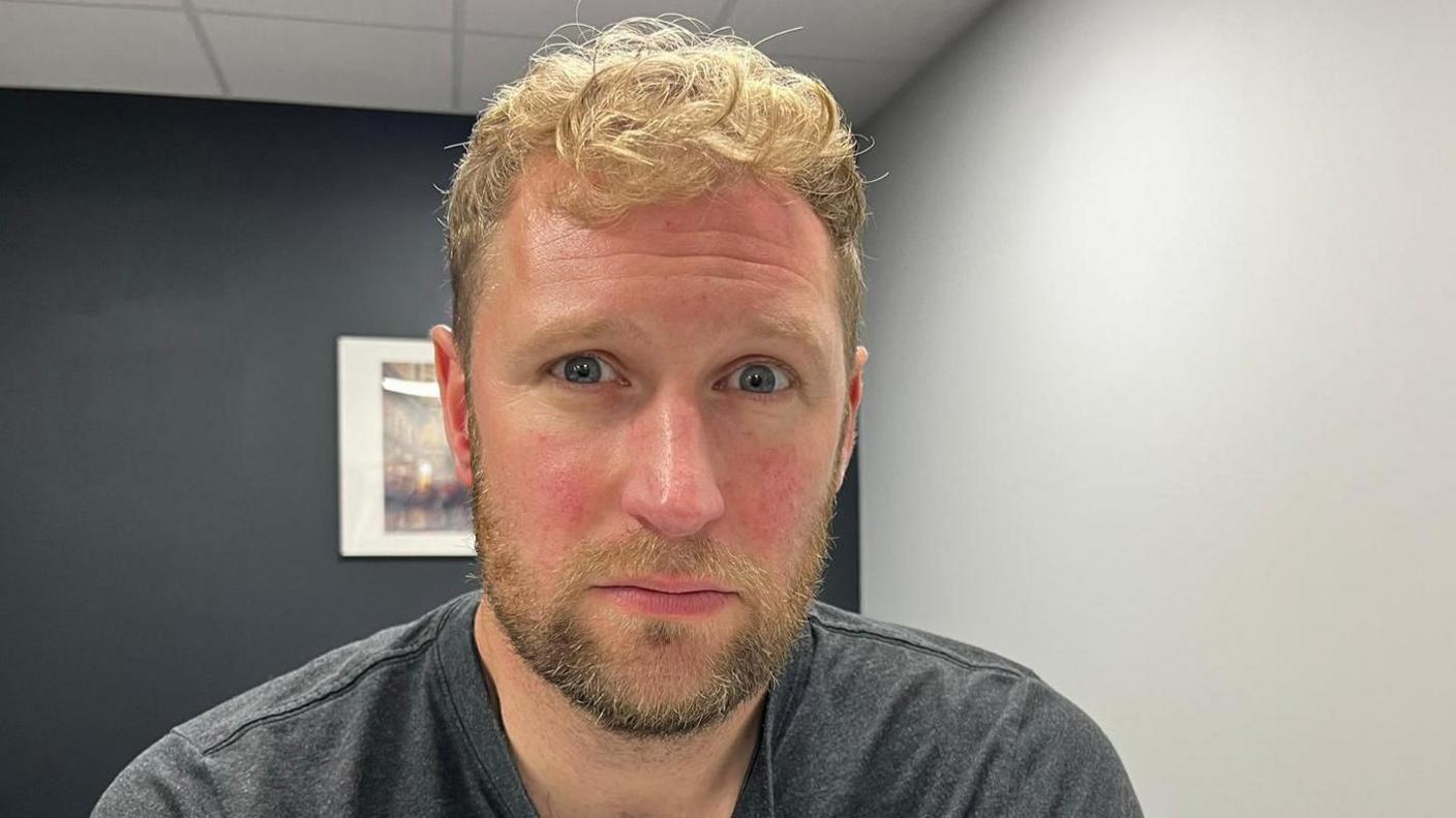 Andy Sirel looking straight at the camera in an office room with dark grey and white walls. He has short curly hair and a beard.