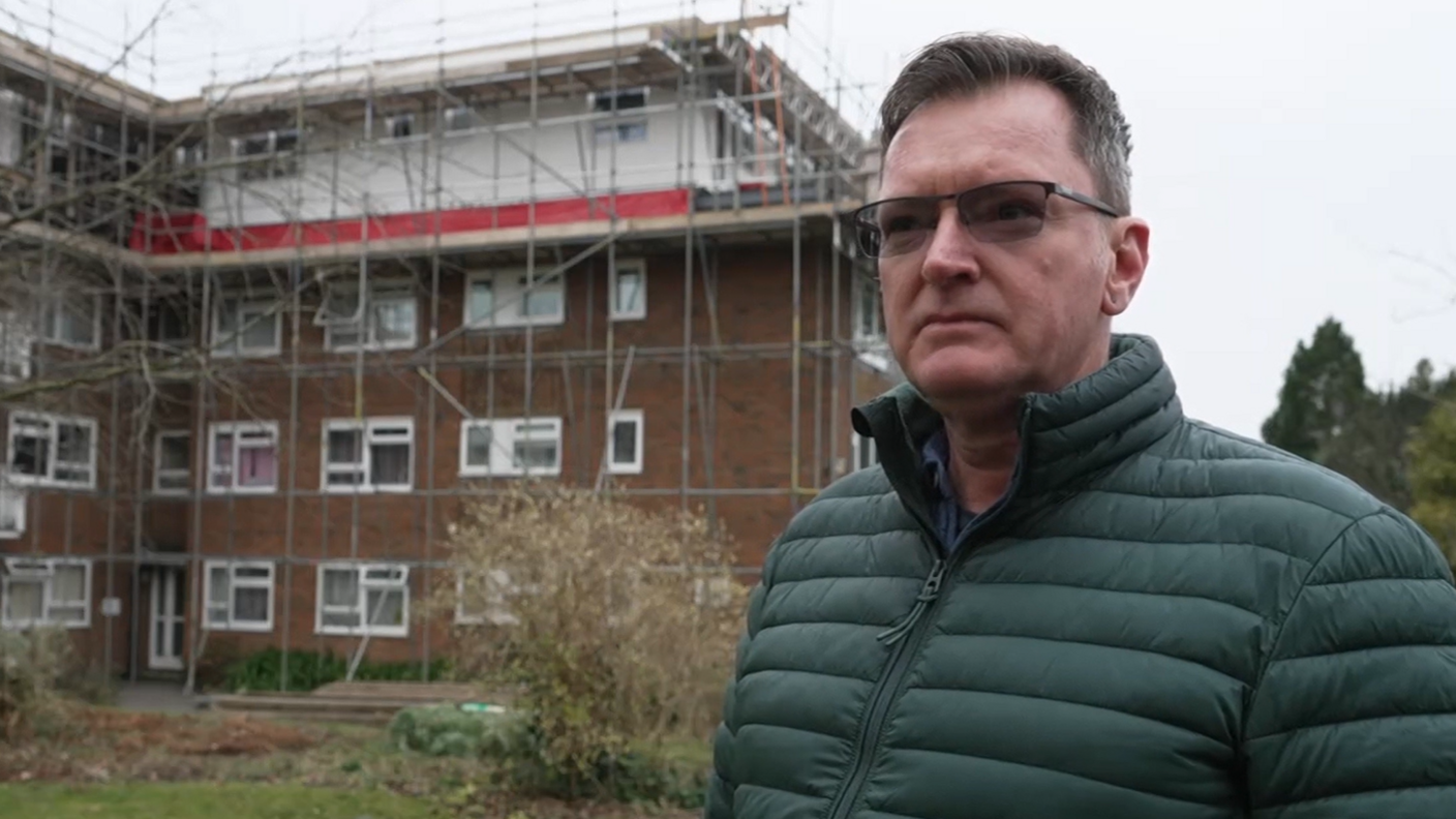 A man in a green coat wearing glasses, with short black hair. He is stood in front of a building wrapped in scaffolding