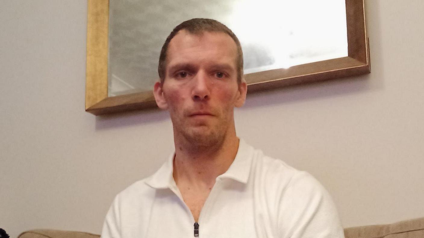 Leigh Walker has short dark hair and beard. He is sitting in a poorly lit room on a cream sofa. Behind him is a wood framed mirror. He is wearing a white polo shirt with a zip front. 