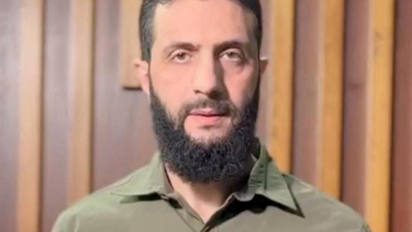 Abu Mohammed al-Jolani looks directly at the camera in this screenshot of a video. He's dressed in a green military shirt and has a dark beard.