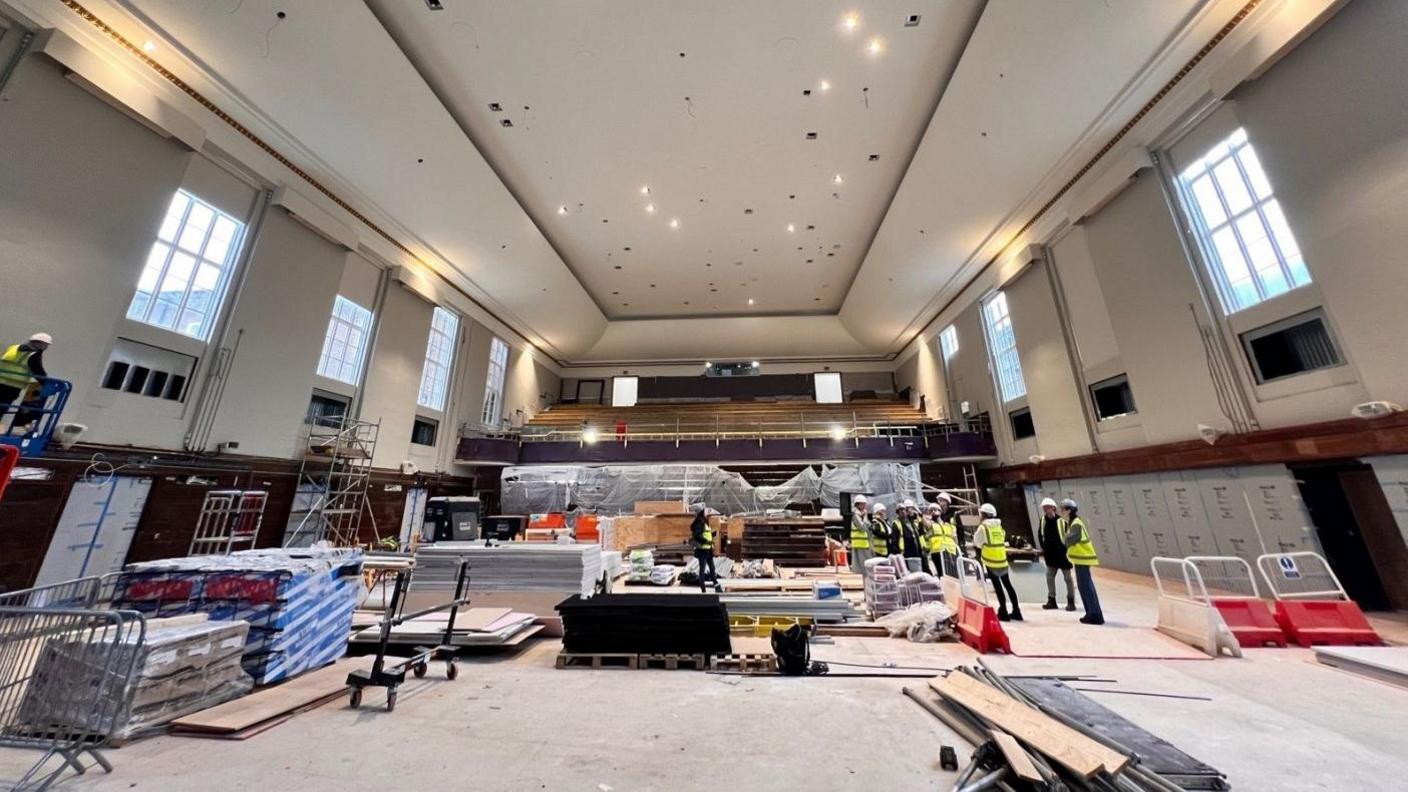 The inside of an arts venue, with people in high-vis jackets, standing in the building with art hats on. There is equipment all around them and building materials. There are racked seats behind them.