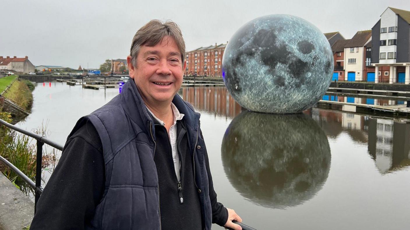 A man stands on the edge of the docks in Bridgwater while behind him the Luke Jerram artwork Fallen Moon can be seen floating on the water