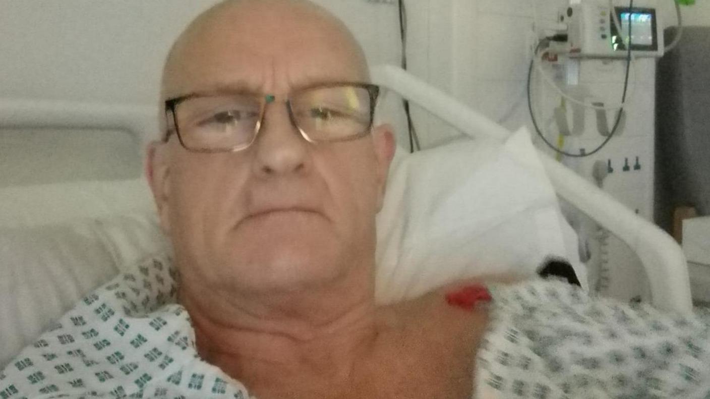 Picture of Steven Richardson in hospital. He is looking at the camera and has a white hospital gown on which has a dark green pattern. His hair is cropped short and he is wearing glasses.