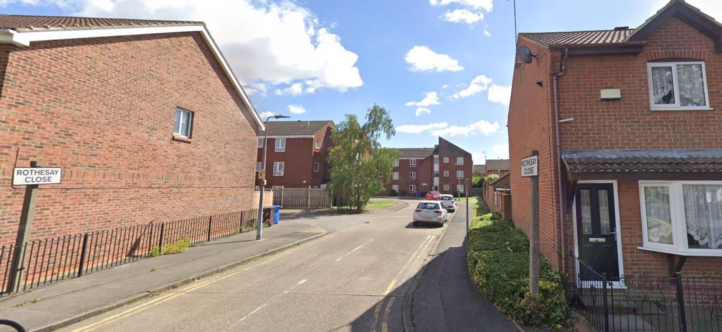 A Google street view image of a residential street with Rothesay Close road name on either side of the entrance to street. On the right is a two-storey redbrick house with a dark green door. On the left is a side wall of a redbrick house with a blue bin in the rear. In the far distance are two blocks of flats. 