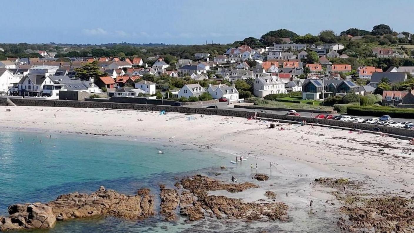 Guernsey with the sea in the corner and a beach. There are houses lined up nearby and a car park with cars parked next to the sand. 
