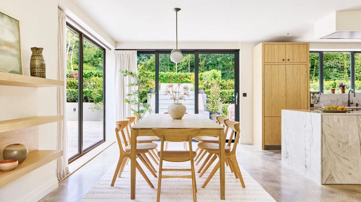 The inside of the house has lots of natural light, a wooden table is in the middle of the room and a marble counter is on the right leading into the kitchen. Through glass windows bushes and the garden are visible.