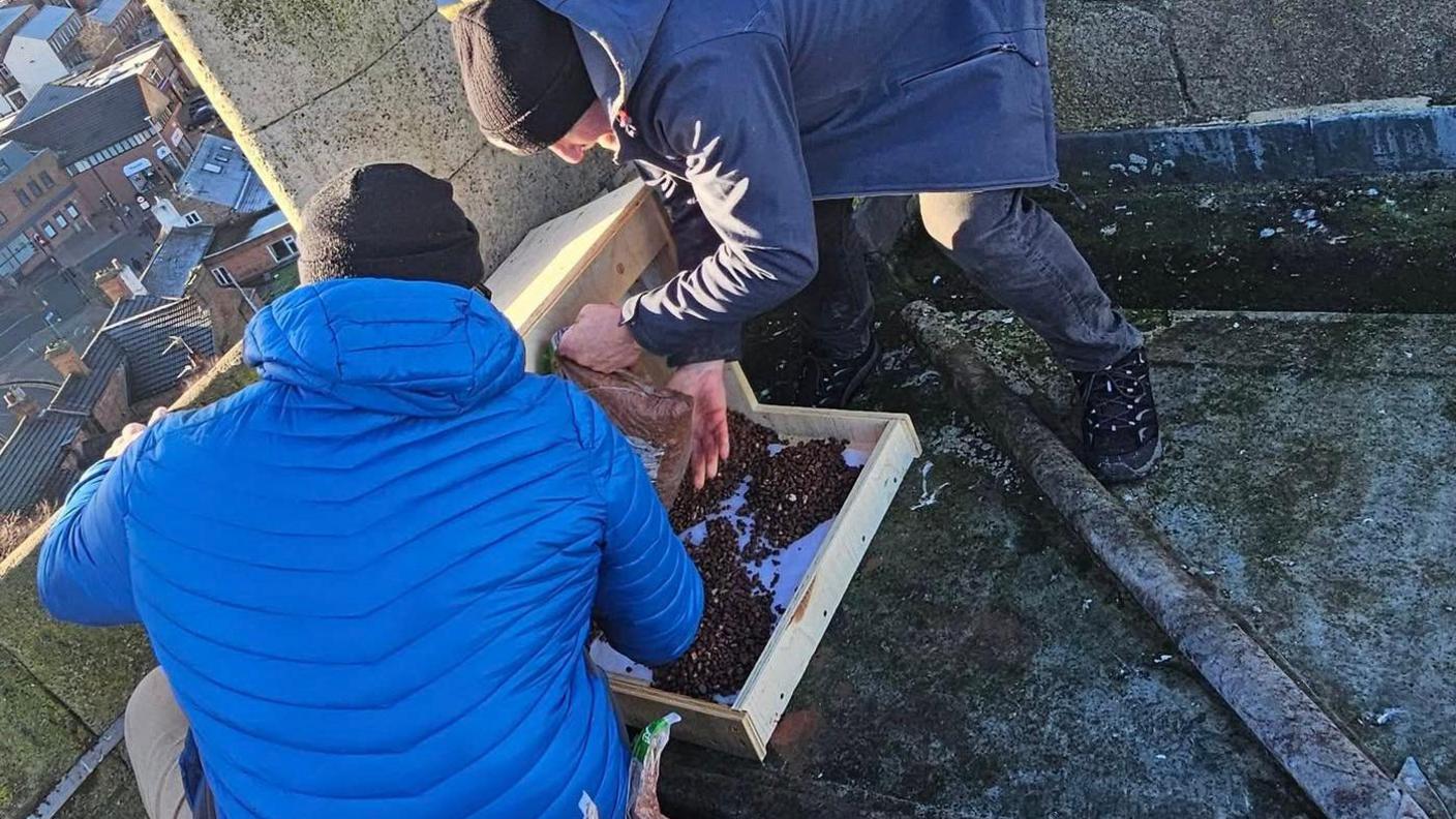 Two men pour gravel into a wooden shelter on the roof with a view to Loughborough below