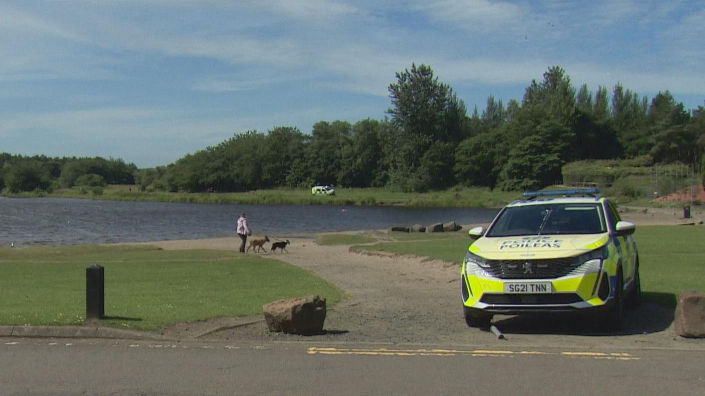 a police car is parked next to the loch. A police officer and a woman walking two dogs are on a path