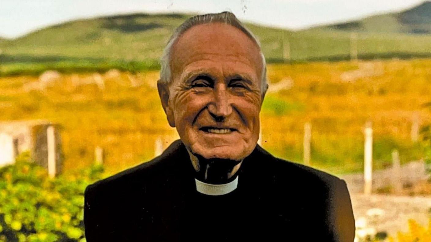 Fr John Morrison pictured when he was an older man. He is wearing a black jacket and a dog collar. There are fields and hills behind him.