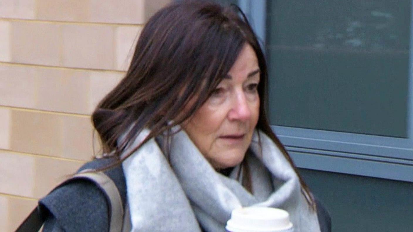 A woman with dark straight hair. She looks cold, and is wearing a grey designer scarf. She is carrying a costa cup. 