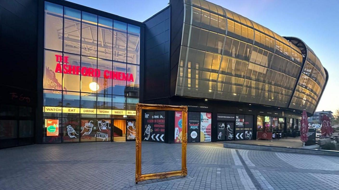 The front entrance to The Ashford Cinema illuminated by lights as the sun sets. A gold picture frame is positioned outside the building's foyer.